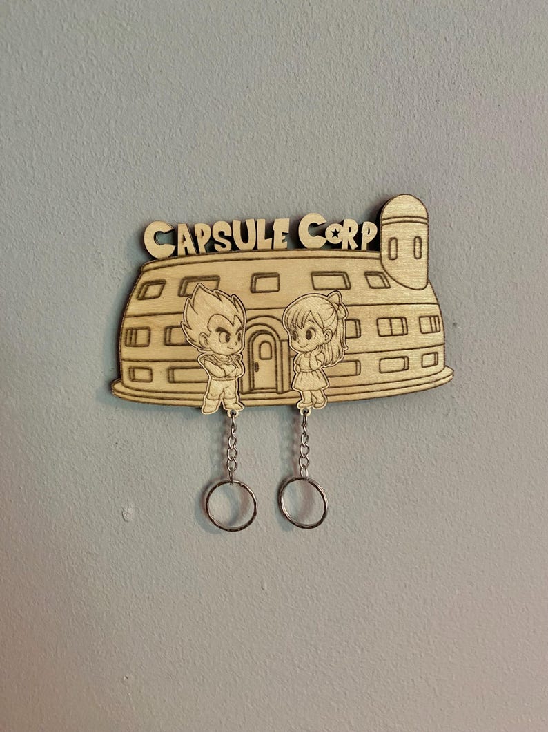May include: A light-colored wooden wall-mounted key holder shaped like the Capsule Corp building. It features engraved anime-style characters and the text "CAPSULE CORP" at the top. Two silver metal chains with key rings hang from the bottom.
