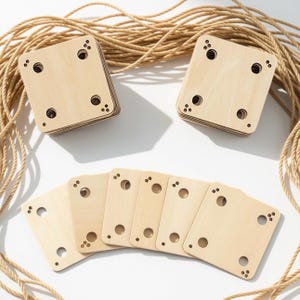 Puede incluir: Dos pilas de piezas cuadradas de madera clara con esquinas redondeadas, cada una con cuatro orificios circulares y pequeños patrones de puntos decorativos en las esquinas, dispuestas junto a seis piezas individuales y una cuerda beige enrollada.