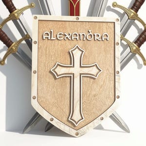 May include: A light-coloured wooden shield featuring the name ALEXANDRA in raised lettering above a central cross design. The shield is bordered by a frame with visible circular rivets and is positioned in front of three crossed metal swords.