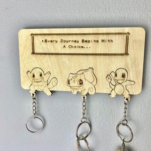 May include: Wooden key holder with three hooks shaped like the Pokemon characters Bulbasaur, Charmander, and Squirtle. The key holder has the text "Every Journey Begins With A Choice..." above the Pokemon characters.