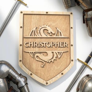 May include: A light-coloured wooden shield featuring a laser-cut dragon design and the name CHRISTOPHER engraved in the centre. The shield has a border with circular cutouts and is surrounded by metal swords and pieces of plate armour.