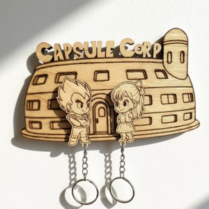 May include: A light-colored wooden wall-mounted key holder featuring the Capsule Corp building design with the text CAPSULE CORP at the top. Two detachable wooden character keychains are suspended from the base by metal chains and rings.