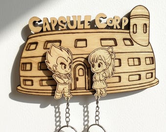 Puede incluir: Un soporte de pared para llaves de madera clara con el diseño del edificio Capsule Corp y el texto CAPSULE CORP en la parte superior. Dos llaveros de madera extraíbles con figuras cuelgan de la base mediante cadenas y anillas de metal.