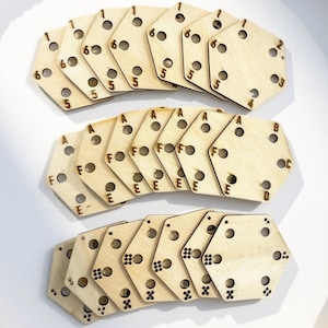 May include: A collection of light-colored wooden hexagonal tiles arranged in three rows. The top row features numbers, the middle row displays letters A through F, and the bottom row shows various dot patterns. Each tile has six circular holes.