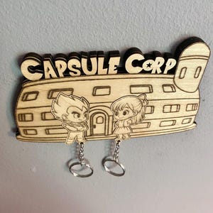 May include: A laser-cut wooden wall-mounted key holder featuring the text CAPSULE CORP and an illustration of a building with two cartoon characters. Two silver metal key rings hang from the bottom of the wooden structure by small chains.