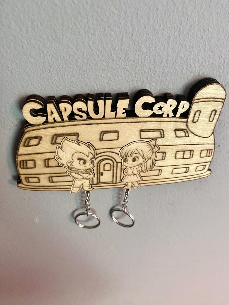 May include: A laser-cut wooden wall-mounted key holder featuring the text CAPSULE CORP and an illustration of a building with two cartoon characters. Two silver metal key rings hang from the bottom of the wooden structure by small chains.