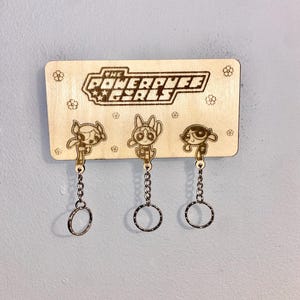 May include: Wooden key holder featuring the Powerpuff Girls logo and three character keychains. The keychains are attached to metal rings with chains. The wooden board has a light brown color with small flower designs.
