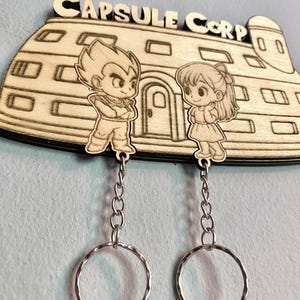 May include: A light-colored wooden wall-mounted key holder featuring the text CAPSULE CORP and an engraved building. Two wooden character cutouts are attached to silver metal chains with circular key rings, depicting a male and female figure.