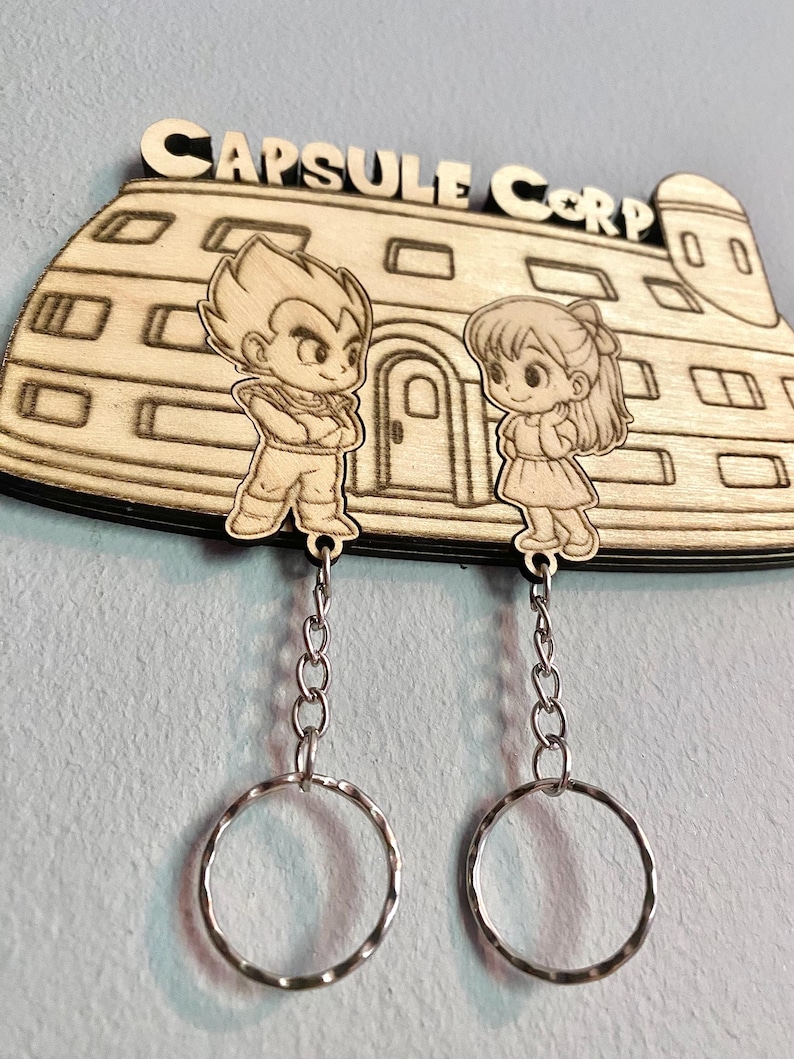 May include: A light-colored wooden wall-mounted key holder featuring the text CAPSULE CORP and an engraved building. Two wooden character cutouts are attached to silver metal chains with circular key rings, depicting a male and female figure.