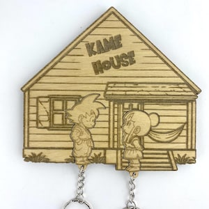 以下が含まれることがあります： 「KAME HOUSE」の文字が入った家のデザインの木製キーホルダー。2人の漫画のキャラクターがキーチェーンに付いています。キーホルダーは明るい茶色の木材で作られており、天然の木目があります。