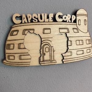 May include: A light-colored wooden wall decoration featuring the text CAPSULE CORP at the top. The design depicts a dome-shaped building with a tower, windows, and a central door, including two silhouette cutouts of characters in the foreground.