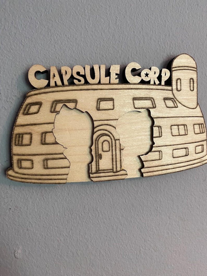 May include: A light-colored wooden wall decoration featuring the text CAPSULE CORP at the top. The design depicts a dome-shaped building with a tower, windows, and a central door, including two silhouette cutouts of characters in the foreground.