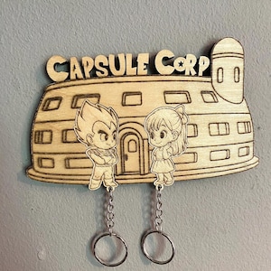 May include: A light-colored wooden wall-mounted key holder featuring a laser-engraved Capsule Corp building design. Two cartoon characters are engraved in front of the building, each attached to a metal chain with a circular key ring at the end.