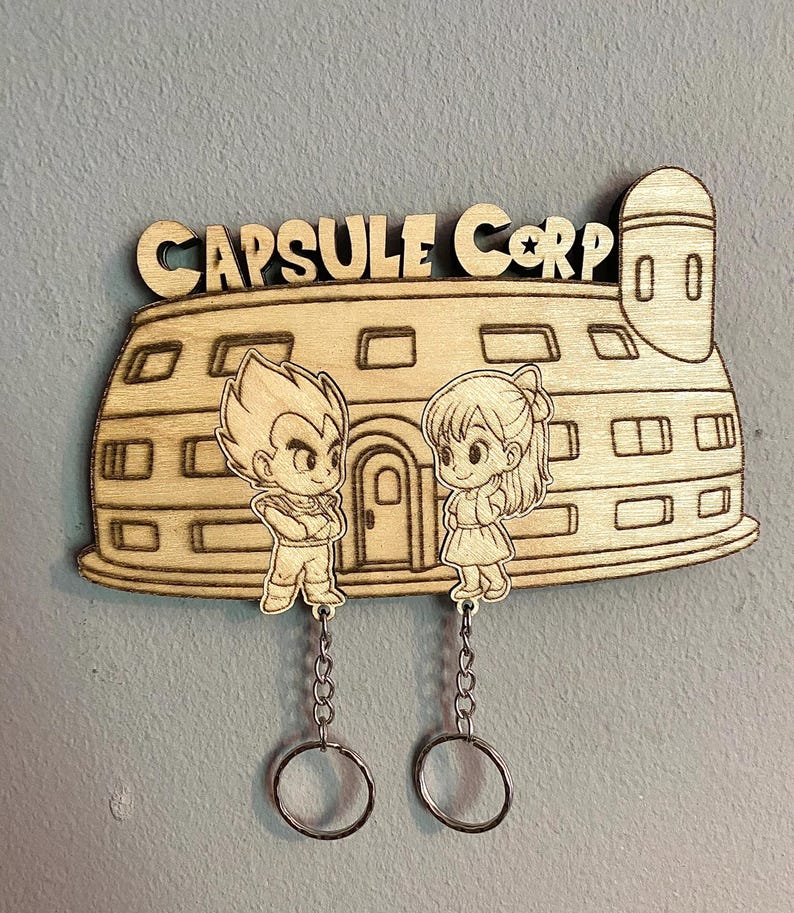 May include: A light-colored wooden wall-mounted key holder featuring a laser-engraved Capsule Corp building design. Two cartoon characters are engraved in front of the building, each attached to a metal chain with a circular key ring at the end.