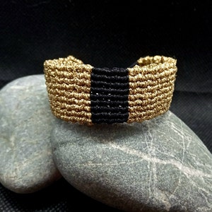 May include: A gold-colored macrame bracelet with a black stripe in the center. The bracelet is wide and has a woven texture. The bracelet is resting on two gray stones against a black background.
