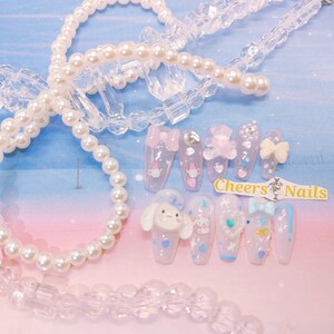 May include: A set of light pink and clear artificial nails with various designs, including bows, hearts, and cartoon characters. The nails are decorated with glitter and small embellishments. The image also includes a pearl necklace and the text "Cheers Nails".