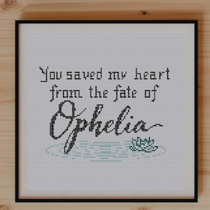 Peut inclure: Une œuvre d'art encadrée au point de croix avec la citation "You saved my heart from the fate of Ophelia". L'œuvre est dans un cadre noir, avec un motif floral en bas. L'image est sur une surface en bois, avec des plantes et des ciseaux.