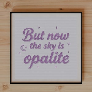 May include: A framed cross-stitch artwork with the text "But now the sky is opalite" in lavender. The artwork is on a wooden surface, with plants, a notebook, and scissors.