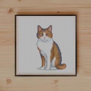 Pode incluir: Uma bordado em ponto cruz emoldurado de um gato com pelo laranja, branco e castanho. A obra de arte é exibida em uma superfície de madeira, juntamente com plantas, um caderno e tesouras.