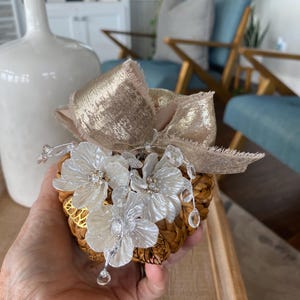 May include: A decorative woven pumpkin ornament featuring a metallic gold ribbon bow, three white pearlescent floral embellishments with central crystal accents, and dangling clear crystal beads attached to the top of the textured brown base.