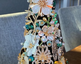 May include: A decorative Christmas tree-shaped ornament, adorned with white flowers, pearls, gold accents, and sparkling jewels. The tree is set against a black background, creating a striking contrast. The top features a crystal finial.