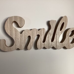 May include: A wooden sign with the word "Smile" carved into it. The sign is made of light brown wood with a natural grain pattern.