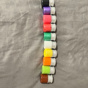 Puede incluir: Un conjunto de 12 pequeños frascos de pintura con tapas plateadas. Los frascos están dispuestos verticalmente en una fila, con los colores que van del negro al marrón. Los colores son negro, morado, rosa, rojo, verde, blanco, amarillo, naranja, verde lima y marrón.