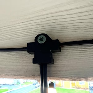 May include: Black plastic string light clips attached to a wooden surface. The clips hold a black wire and a light bulb. The light bulb is lit and has a warm glow. The wooden surface has a light brown color.