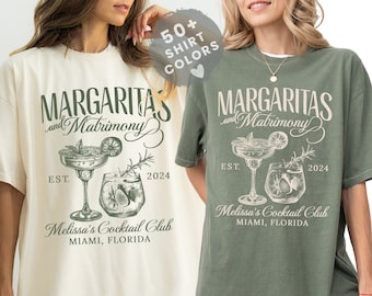Może przedstawiać: Dwie koszulki z nadrukiem graficznym dwóch szklanek z margaritą i tekstem "Margaritas and Matrimony" z rokiem 2024 i "Melissa's Cocktail Club Miami, Florida". Koszulki są w kolorze kości słoniowej i zielonym mchu.