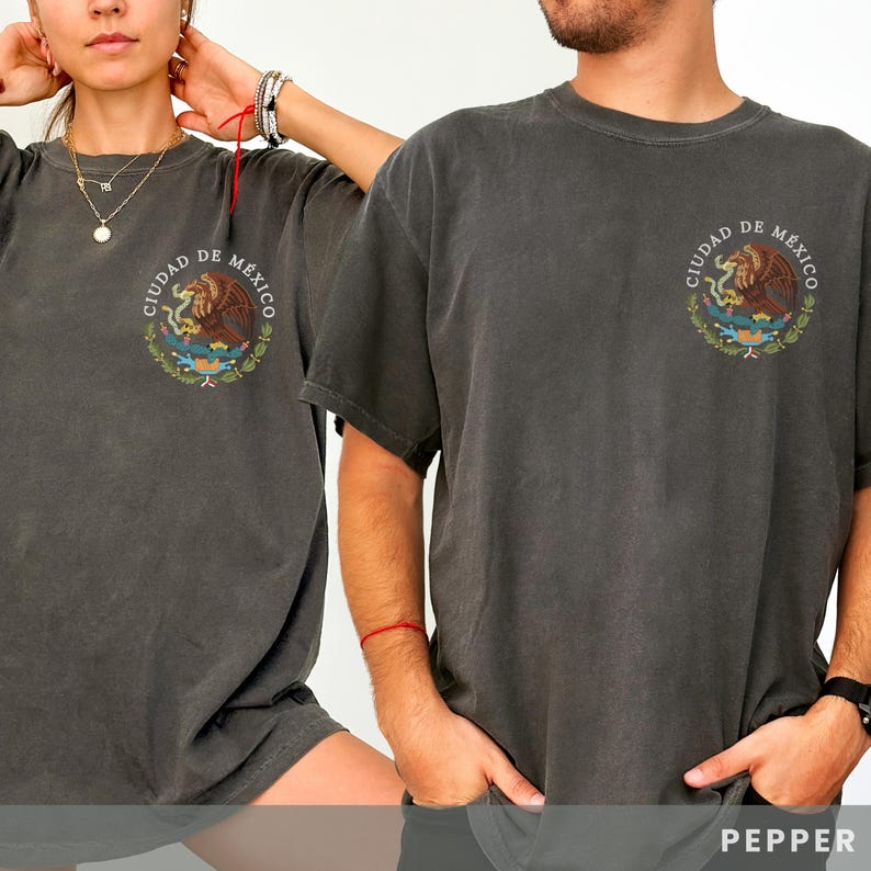 May include: Two people wearing charcoal grey t-shirts with a vintage-style graphic of the Coat of Arms of Mexico City. The graphic features the text "CIUDAD DE MEXICO" and an eagle perched on a cactus.
