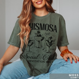 May include: A green t-shirt with a black graphic print of three women in cocktail glasses. The text on the shirt reads "Mimososa Social Club Invitation Only" and "1983".