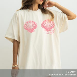 Pu&ograve; includere: T-shirt color avorio con due grafiche di conchiglie rosa. Il testo "Cabo San Lucas, May 2020" &egrave; stampato sotto la conchiglia destra. La parte inferiore della maglietta ha il testo "IVORY CLR#18 WATERMELON".