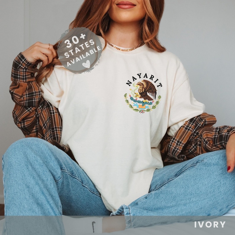A Model in a sitting position with ivory colored comfort colors shirt with brown graphic of Mexican Coat of Arms.