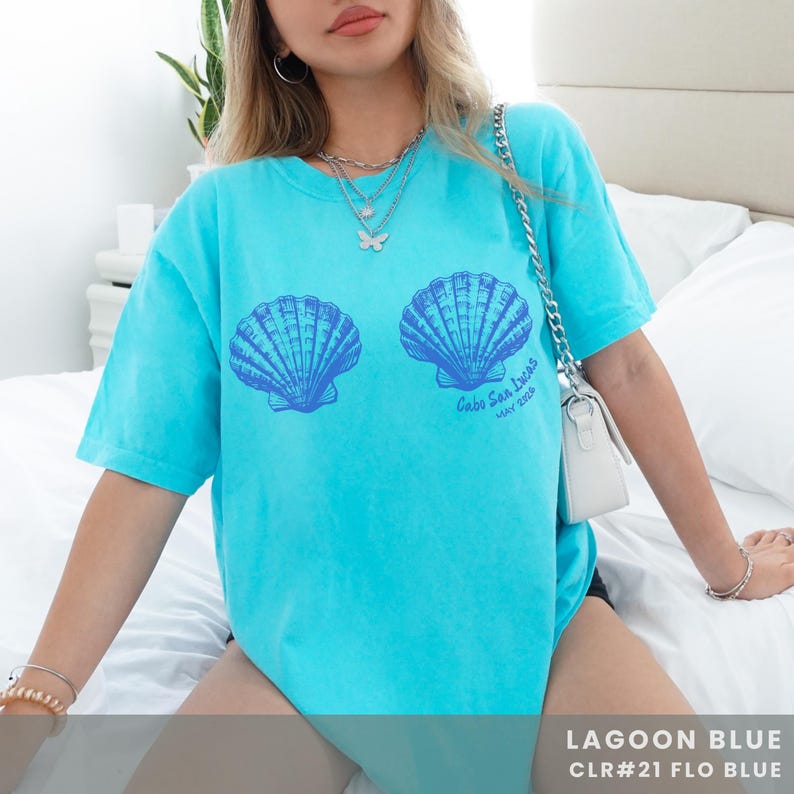 Pu&ograve; includere: T-shirt blu laguna con due grafiche di conchiglie blu. Il testo "Cabo San Lucas MAY 2023" &egrave; stampato sotto le conchiglie. La parte inferiore della maglietta riporta la scritta "LAGOON BLUE CLR#21 FLO BLUE".