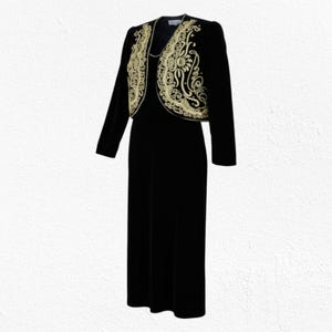 Può includere: Un abito in velluto nero con bolero abbinato. La giacca presenta ricami dorati intricati lungo i bordi e lo scollo. L'abito è un design semplice ed elegante, adatto alle occasioni formali.