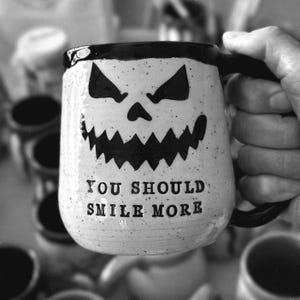 A handmade pottery mug made from white speckled clay that reads “YOU SHOULD SMILE MORE”. The inside, rim, and handle are glazed obsidian black. This is a black and white photo.