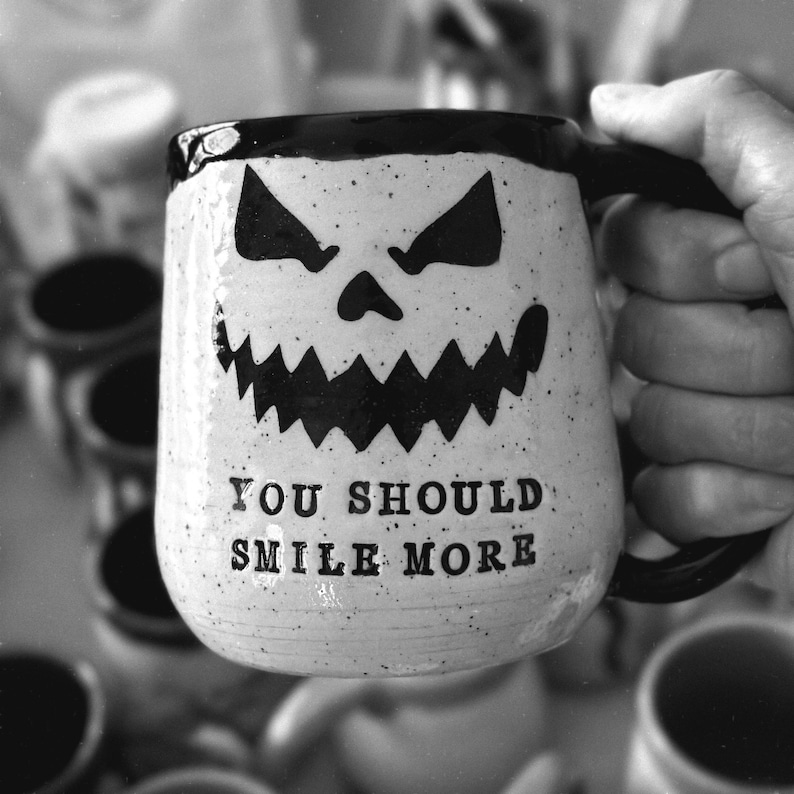 A handmade pottery mug made from white speckled clay that reads “YOU SHOULD SMILE MORE”. The inside, rim, and handle are glazed obsidian black. This is a black and white photo.