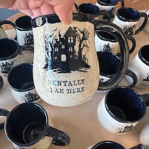 May include: A collection of white ceramic mugs with black accents. The mugs feature a silhouette of a haunted house and the text "Mentally I am here".