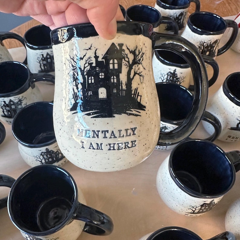May include: A collection of white ceramic mugs with black accents. The mugs feature a silhouette of a haunted house and the text "Mentally I am here".