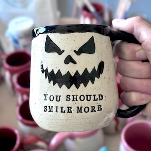 May include: A ceramic mug with a black handle and rim, featuring a spooky jack-o'-lantern face design. The mug has the text "YOU SHOULD SMILE MORE" printed below the face. The mug is cream-colored with black speckles.