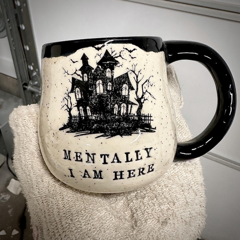 A mitted hand holding up a white speckled clay mug that reads “MENTALLY I AM HERE” in black letters with a black image of a haunted house. The interior, handle, and rim are glazed glossy black.