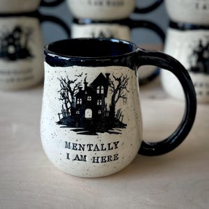 A white speckled clay handmade pottery mug that reads “MENTALLY I AM HERE” in black letters with a black image of a haunted house. The interior, handle, and rim are glazed glossy black.