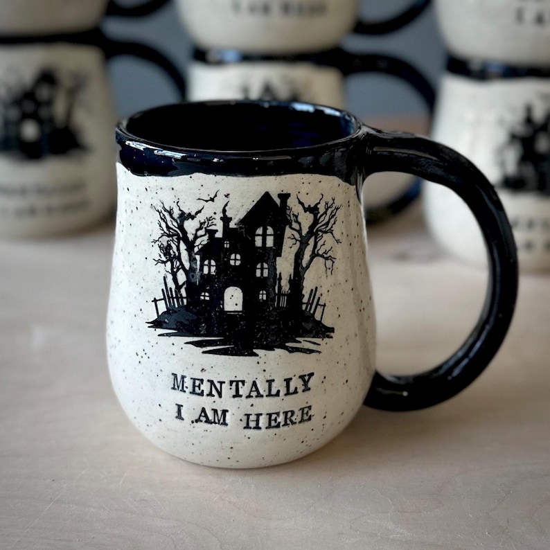 A white speckled clay handmade pottery mug that reads “MENTALLY I AM HERE” in black letters with a black image of a haunted house. The interior, handle, and rim are glazed glossy black.