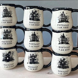 May include: A stack of eight white ceramic mugs with black rims and handles. Each mug features a black silhouette of a spooky house with trees and the text "Mentally I am here".