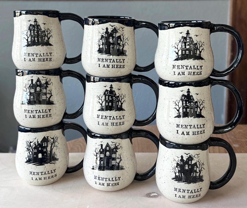 May include: A stack of eight white ceramic mugs with black rims and handles. Each mug features a black silhouette of a spooky house with trees and the text "Mentally I am here".