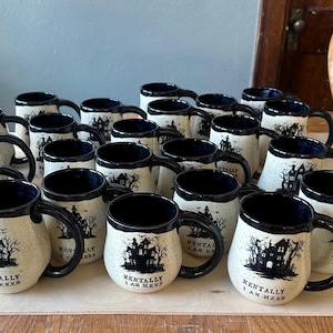 May include: A collection of white ceramic mugs with black accents and a spooky house design. The mugs have the text "Mentally I am here" printed on them.