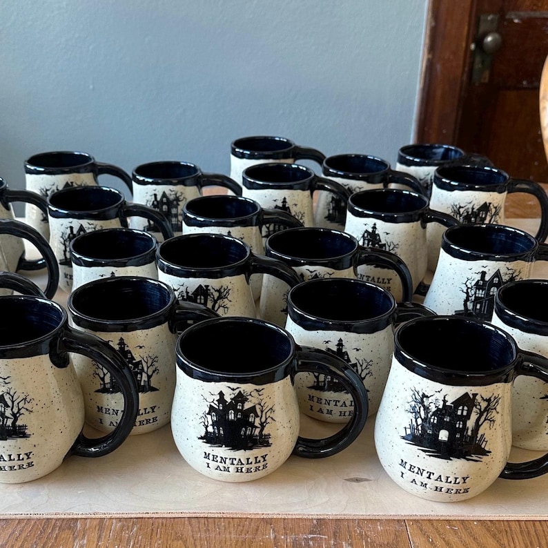 May include: A collection of white ceramic mugs with black accents and a spooky house design. The mugs have the text "Mentally I am here" printed on them.