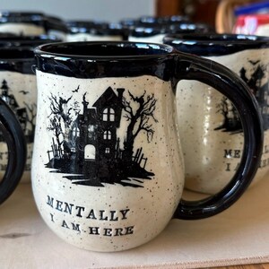 May include: A white ceramic mug with a black silhouette of a haunted house and trees. The mug has a black rim and handle. The text "Mentally I am here" is printed on the front of the mug.