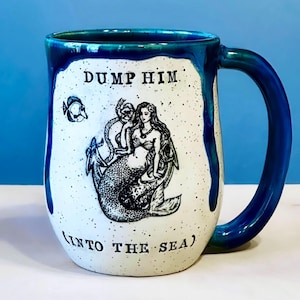 May include: White ceramic mug with a blue handle and rim. The mug features a black and white illustration of a mermaid and the text "DUMP HIM (INTO THE SEA)". A small fish is also depicted on the mug.