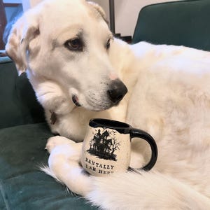 Cassio, a 100 lb. akbash dog, curled up with a mug. The mug is a white speckled clay mug that reads “MENTALLY I AM HERE” in black letters with a black image of a haunted house. The interior, handle, and rim are glazed glossy black.